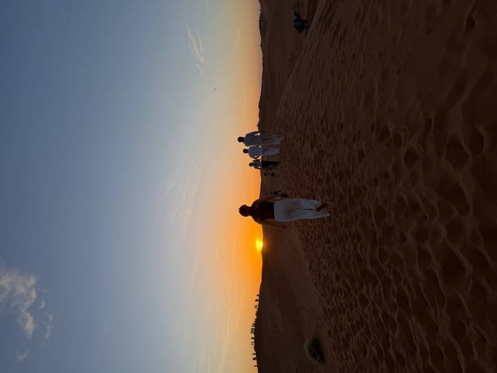 Tourists climb a sandy ridge toward a colourful sunset in the Sahara desert.