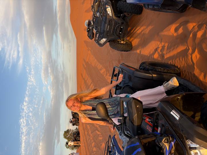 Smiling woman sits on a quad bike with dramatic orange dunes and long evening shadows behind.