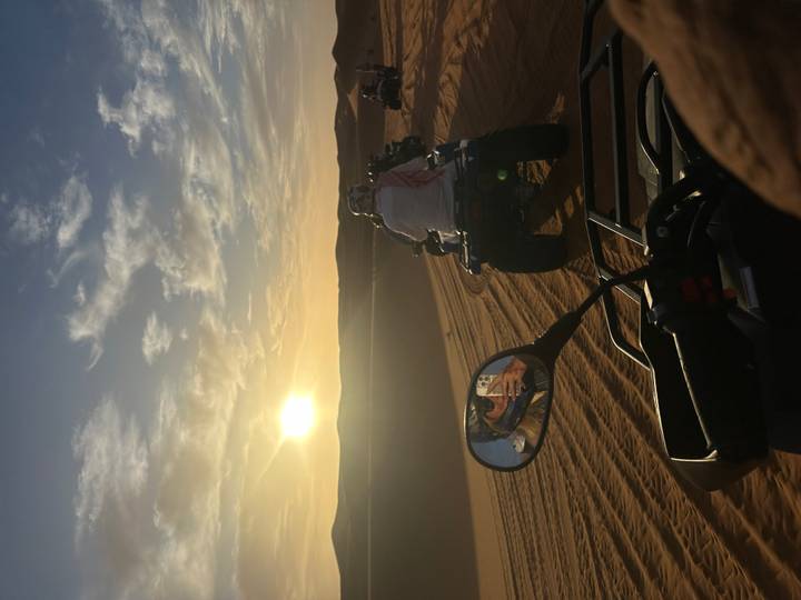 Line of quad bikers drives over soft desert sand toward the setting sun.