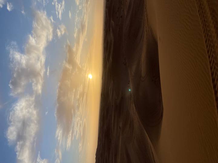 Golden sun hangs low over vast rippling Sahara dunes under a partly cloudy sky.