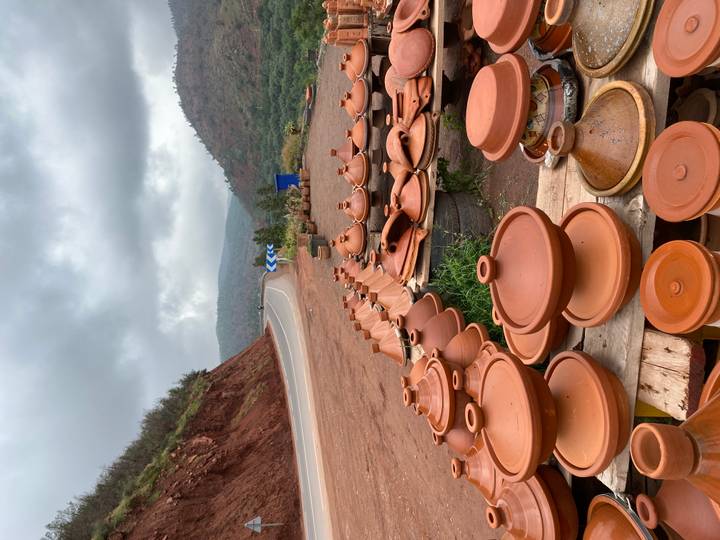 Stacks of terracotta tagine pots line a roadside stall with winding mountain highway in background.