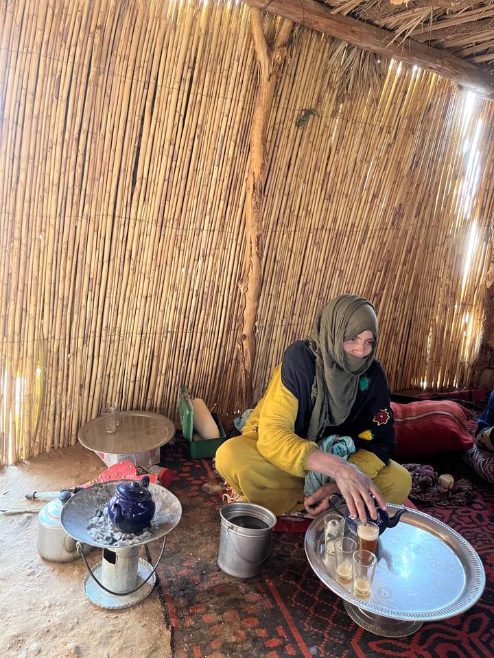 A young woman in traditional dress sits inside a simple reed hut, soft light illuminating the rustic interior.