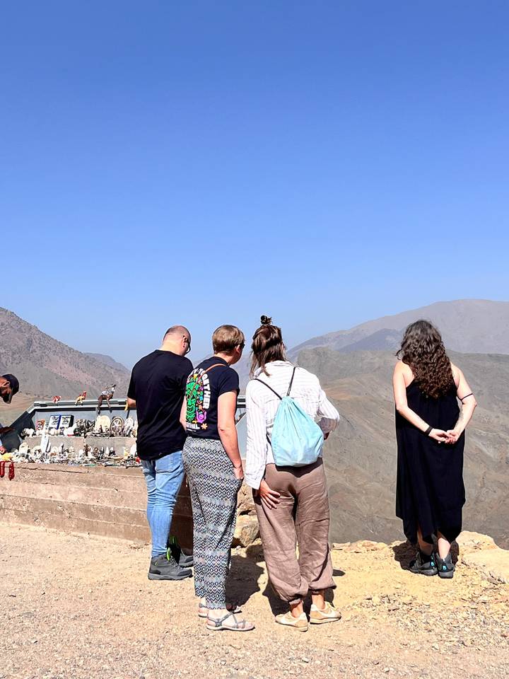 Travellers stand at a mountain viewpoint, backs to camera, admiring the Atlas range and roadside shrine.