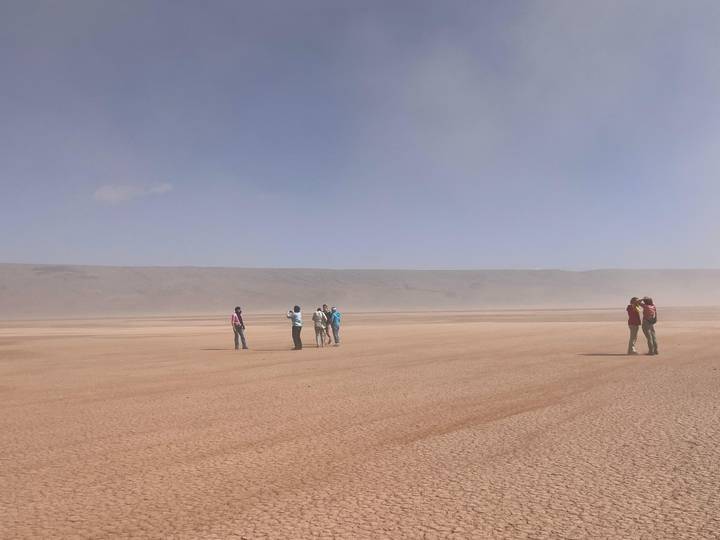 Small groups of travellers stand on a vast, dusty clay pan under a hazy blue sky.