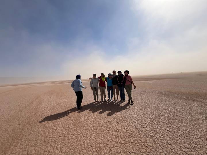 Guide gathers a group for a photo on a cracked salt flat while dust swirls in distant haze.