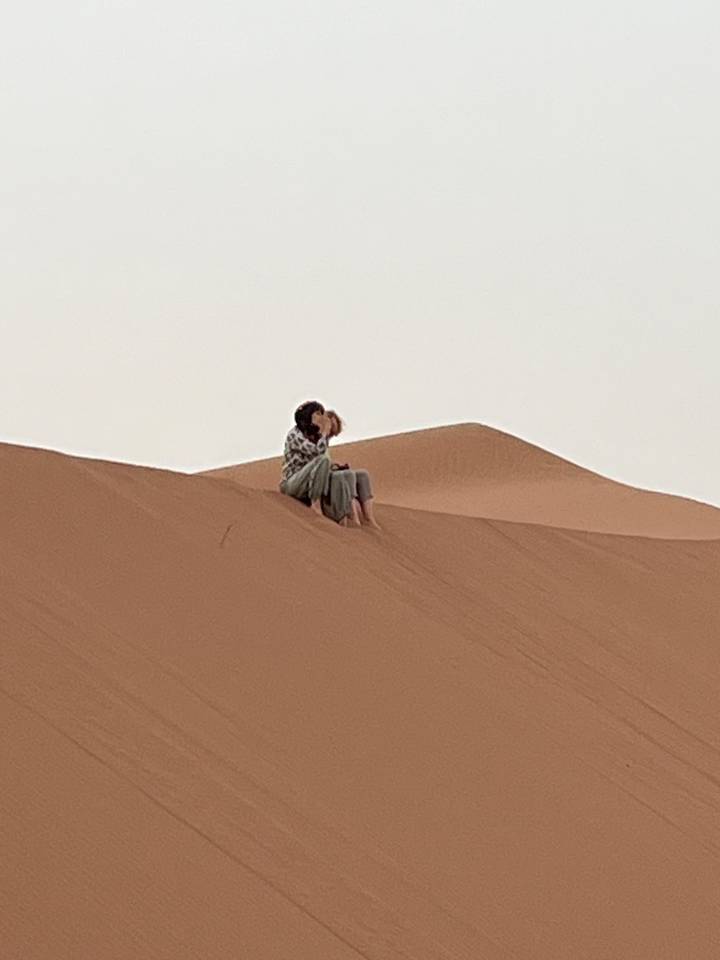 Romantic couple sits on the crest of an orange sand dune sharing a quiet kiss.