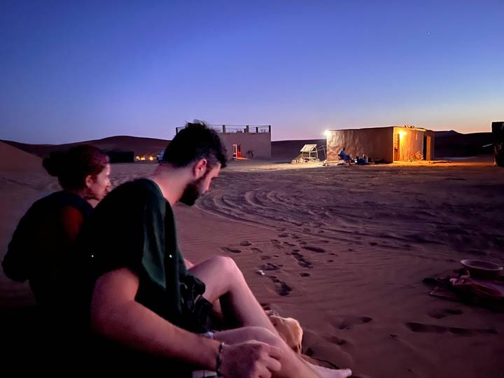 Couple sits near a small desert camp at dusk with warm lamplight glowing from a clay hut.