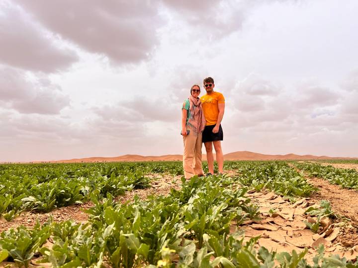 Young couple standing in a green crop field with Sahara sand-dune ridges in the distance under cloudy skies.