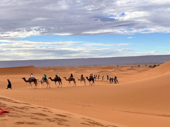 Long camel caravan led by a guide traverses golden Sahara dunes beneath a patchy sky.