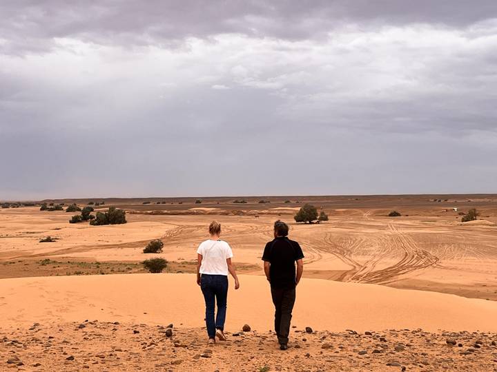 Two travelers stand atop a low sand dune looking over wide tracks and scattered shrubs in the desert.
