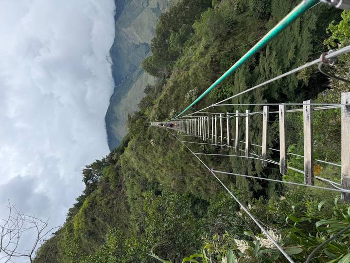 Narrow hanging bridge made of wooden planks and cables stretches across a deep green gorge toward distant hills.