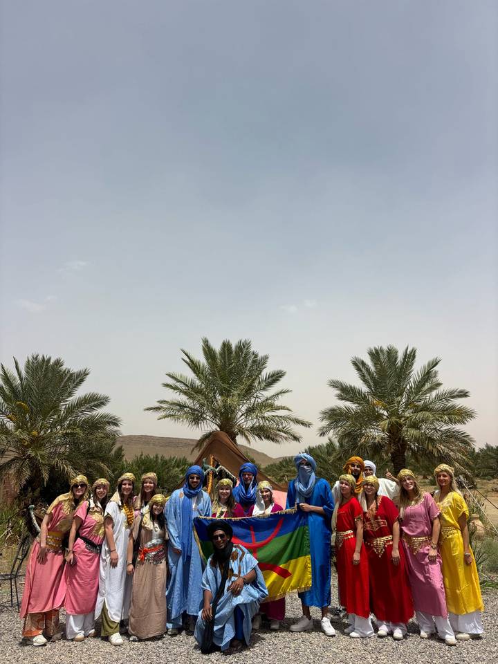 Palm trees and a small desert tent with a few people in colorful turbans beneath a wide pale sky.
