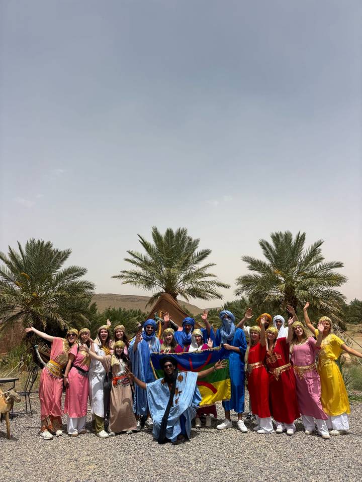 A cheerful group in bright Berber head-scarves poses under palm trees at a desert camp.