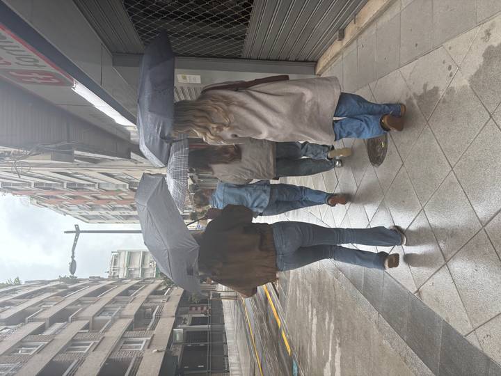 Women with umbrellas walk down a wet sidewalk lined by tall buildings.
