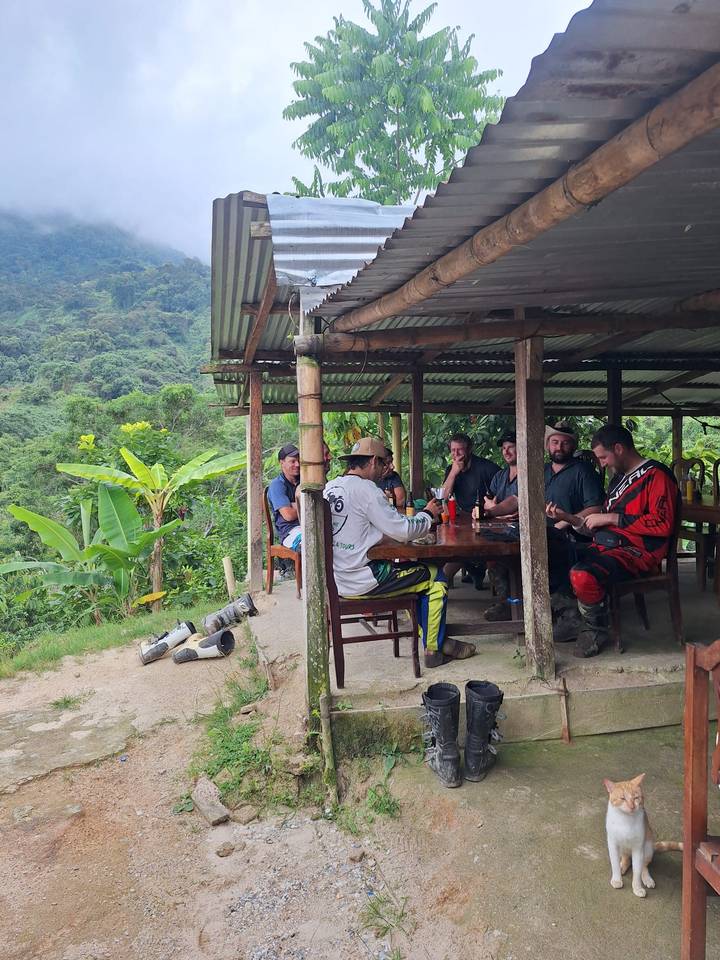 Riders relax around a wooden table at a rustic jungle shelter with lush green valley backdrop.