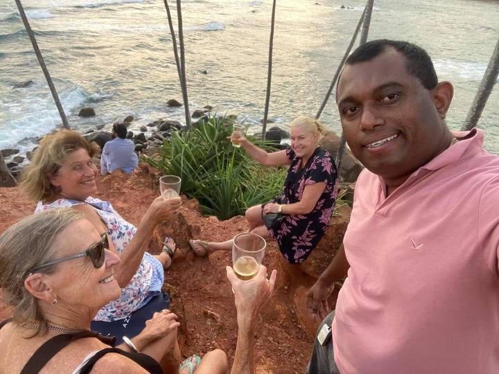 Selfie of travellers raising drinks on a red-rock cliff above crashing ocean waves at dusk.