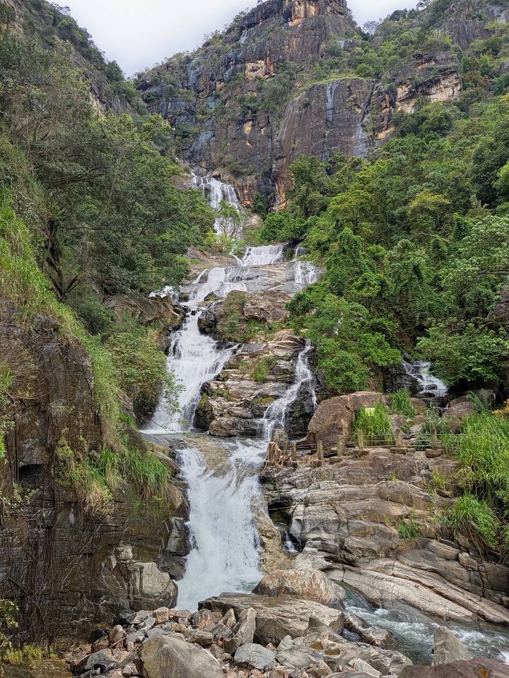 Multi-tiered waterfall cascades through dense green jungle vegetation over rocky ledges.