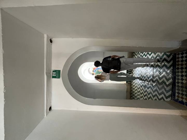Tourists walk down a narrow arched corridor with patterned floor tiles toward daylight.