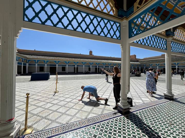Wide courtyard of Bahia Palace with patterned tiles; tourists explore while one person kneels playfully for a photo.