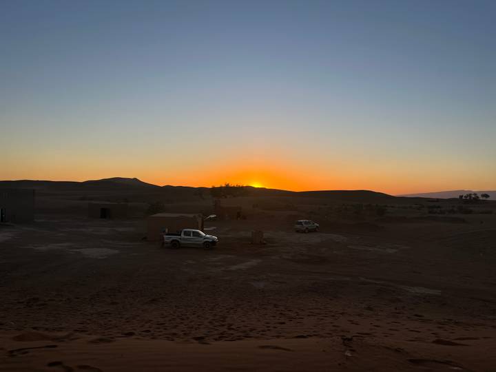 Desert camp with pickup trucks and low buildings silhouetted against an orange sunset over sand plains