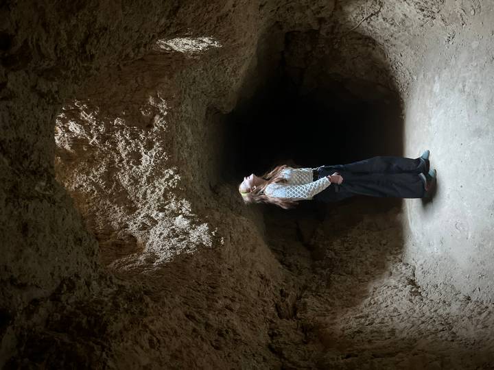 Traveler looks up toward a shaft of sunlight in a narrow earthen cave chamber.