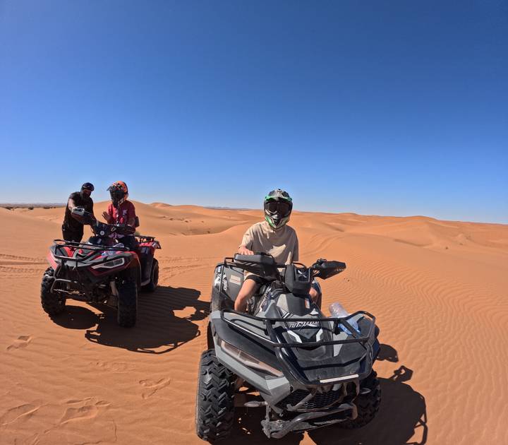 Travelers ride quad bikes across vast orange dunes under a clear blue desert sky.