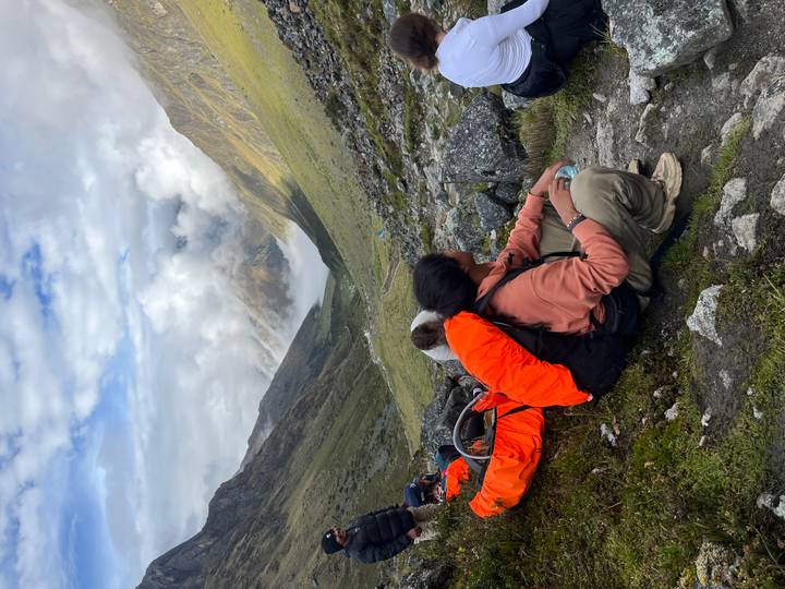 Hikers rest on rocky alpine trail with dramatic clouds engulfing distant Andean valley.