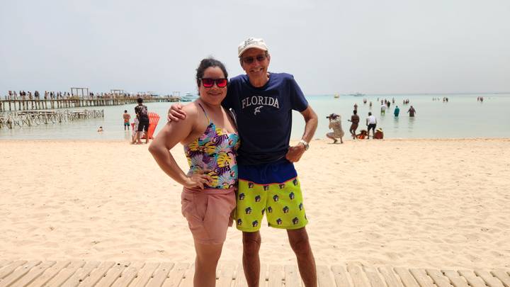 Smiling man and woman pose on a sandy beach beside shallow turquoise water with a pier and bathers in the background.