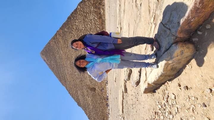 Two women stand on a stone ledge with the Great Pyramid rising sharply behind them against a clear blue sky.