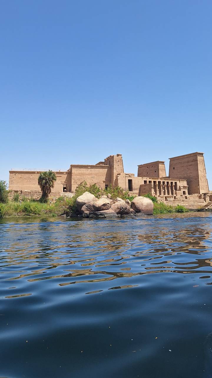 Philae Temple’s sandstone walls rising above the Nile with rocky islet and palms in foreground.