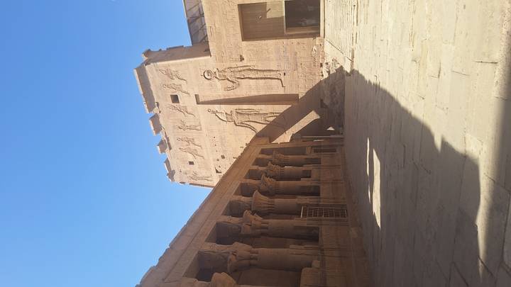 Tall relief-covered pylon and columned courtyard inside an ancient Egyptian temple under clear blue sky.