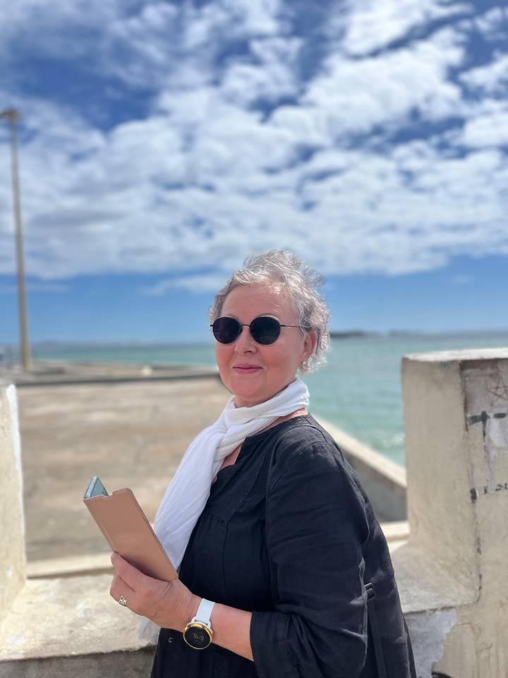 Portrait of a stylish woman with sunglasses and white scarf standing beside a bright turquoise sea under scattered clouds.
