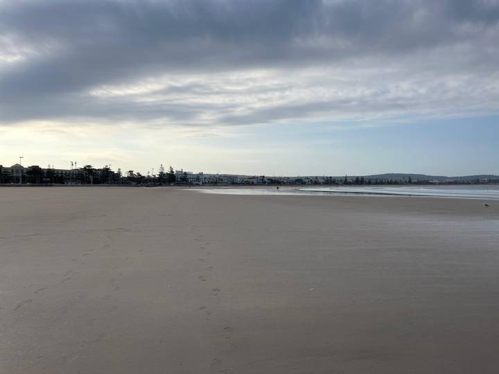 Wide, almost empty sandy beach with distant promenade and soft early morning light.