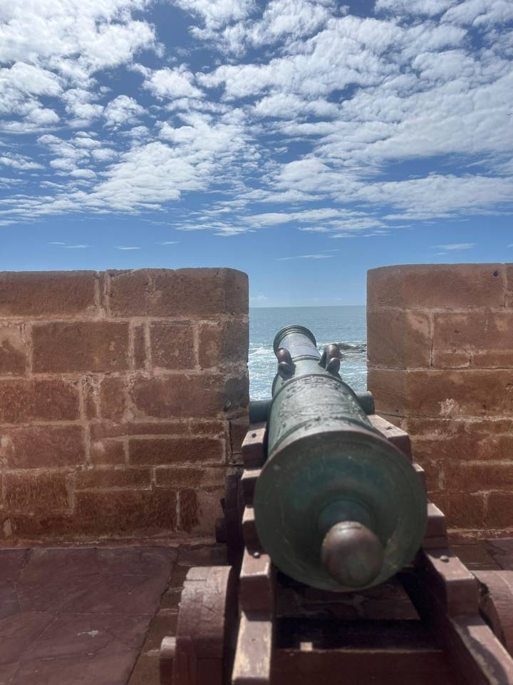 Historic cannon points through stone ramparts toward the Atlantic Ocean under a bright sky.