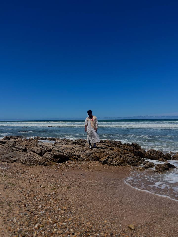 Traveler in flowing robe stands on rocky shore with crashing Atlantic waves and blue sky.
