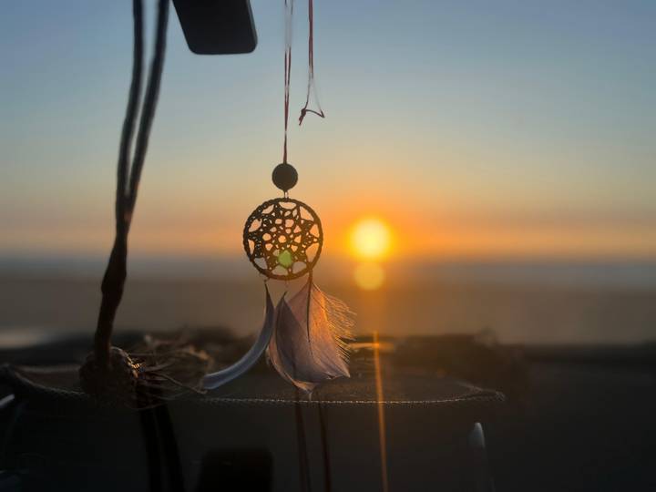 Dreamcatcher silhouette hangs in focus with glowing sunset and blurred shoreline beyond.
