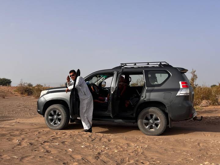 Man in traditional clothing leaning on an SUV parked on sandy desert ground.