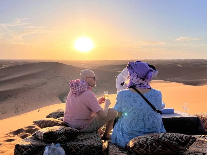 Couple enjoying drinks on cushioned mats while sun sets over vast sand dunes