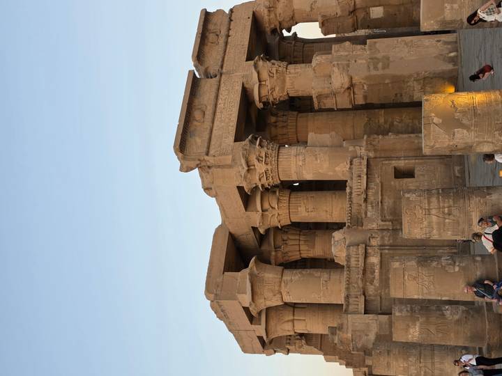 Sand-colored temple with thick columns decorated with hieroglyphs as tourists sit at its base during late afternoon light.