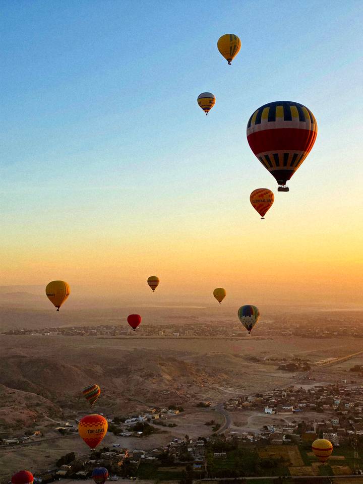 Multiple colourful hot-air balloons floating above a hazy desert cityscape at sunrise.