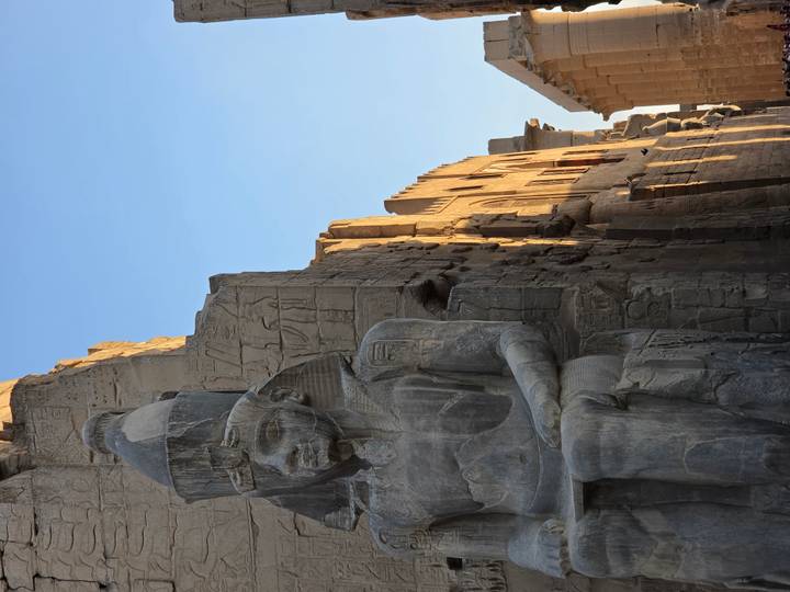 Huge seated pharaoh statue outside temple walls warmly lit by early morning sun with carved hieroglyphs in background.