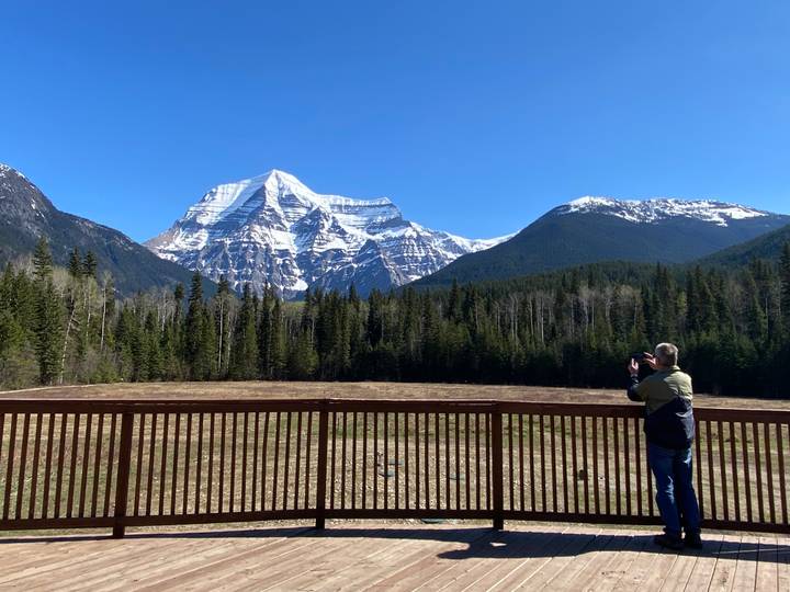 Photographer on a viewing deck capturing the snow-covered peak of Mount Robson rising above dense forest