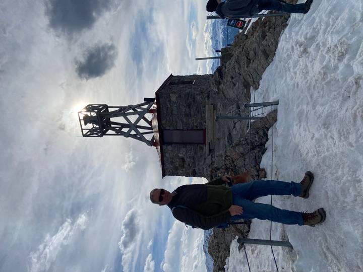 Hiker standing on a snowy summit beside an old stone weather station silhouetted against the sun
