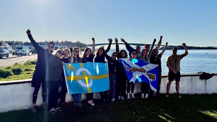 Excited tour group posing with colourful flags beside a coastal inlet under bright sunshine.