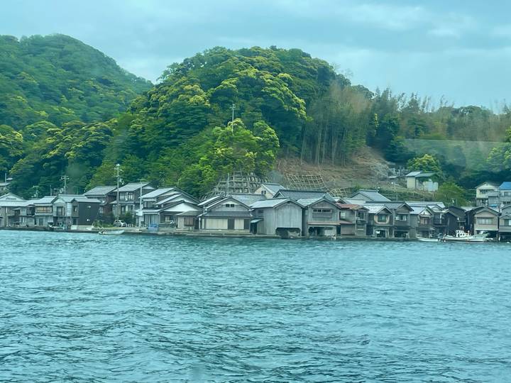 Traditional wooden boathouses lining a quiet coastal village backed by dense green hills.