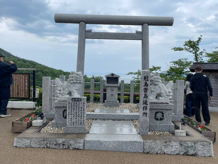 Granite torii gate and shrine monument atop a hill with travellers taking photos and hazy views beyond.
