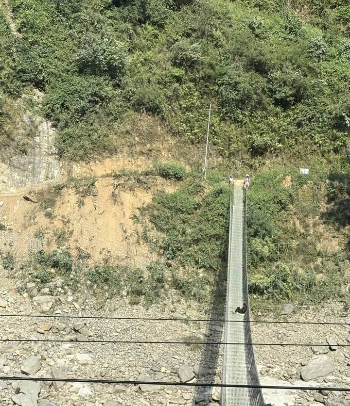 Two trekkers crossing a long narrow suspension bridge over a deep forested ravine.