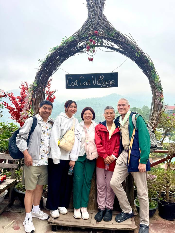 Travellers stand beneath an archway sign reading “Cat Cat Village” with misty hills in the background.