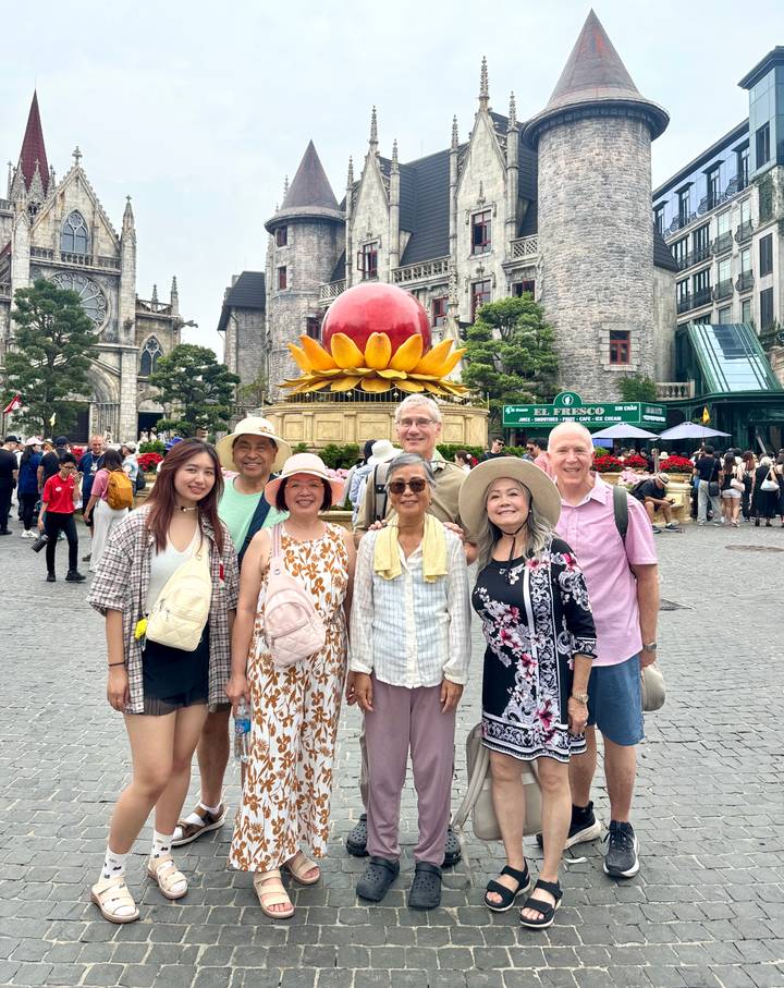 Tour group poses in a lively European-style square with a giant lotus sculpture and crowd behind.