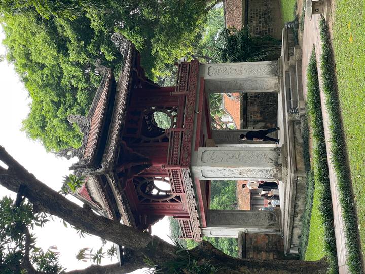 Historic red wooden pavilion with tiled roof and stone pillars set in a leafy garden.